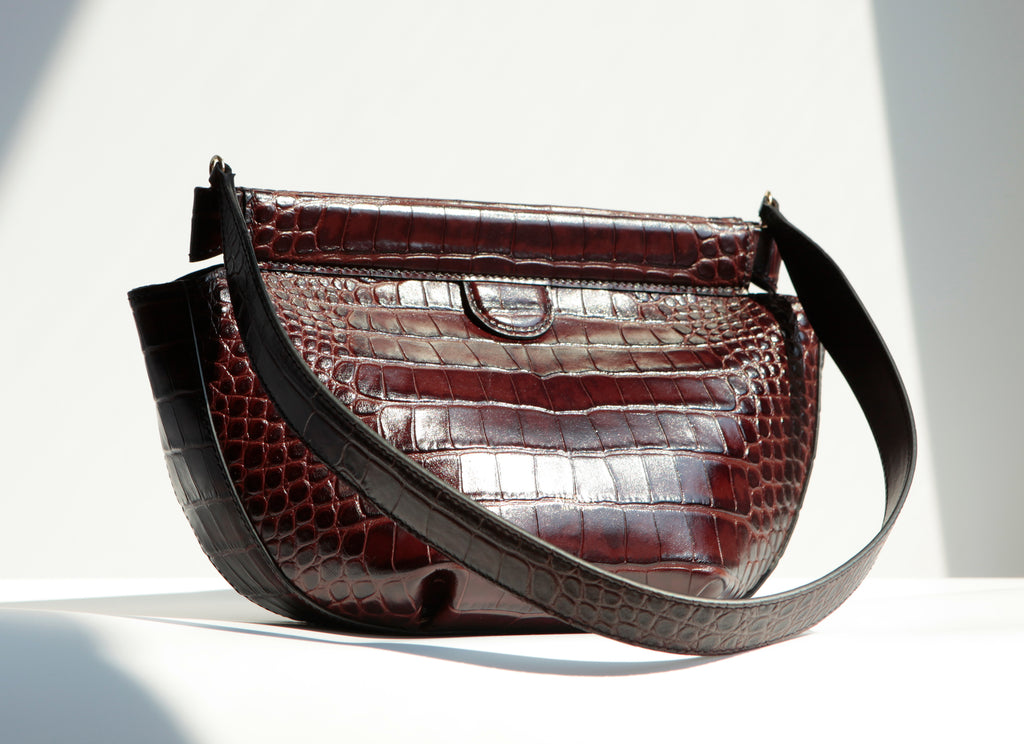 Phoebe Dumpling Shoulder Bag shows the bottom pleat in this three quarter view and croc-embossed texture of the leather.  A pull tab on both sides of the bag allows to open the top hinge bar frame closure.