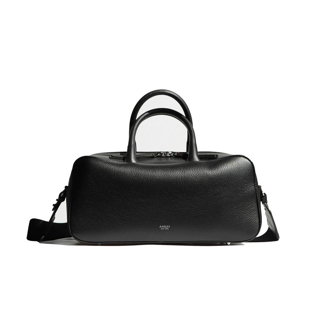 Side View of the Ines Mini Carryall Satchel. Shows the width and top zipper that extends on third way down the sides of the bag to allow for the top to open wide.  Bottom studs to protect the bottom leather corners.