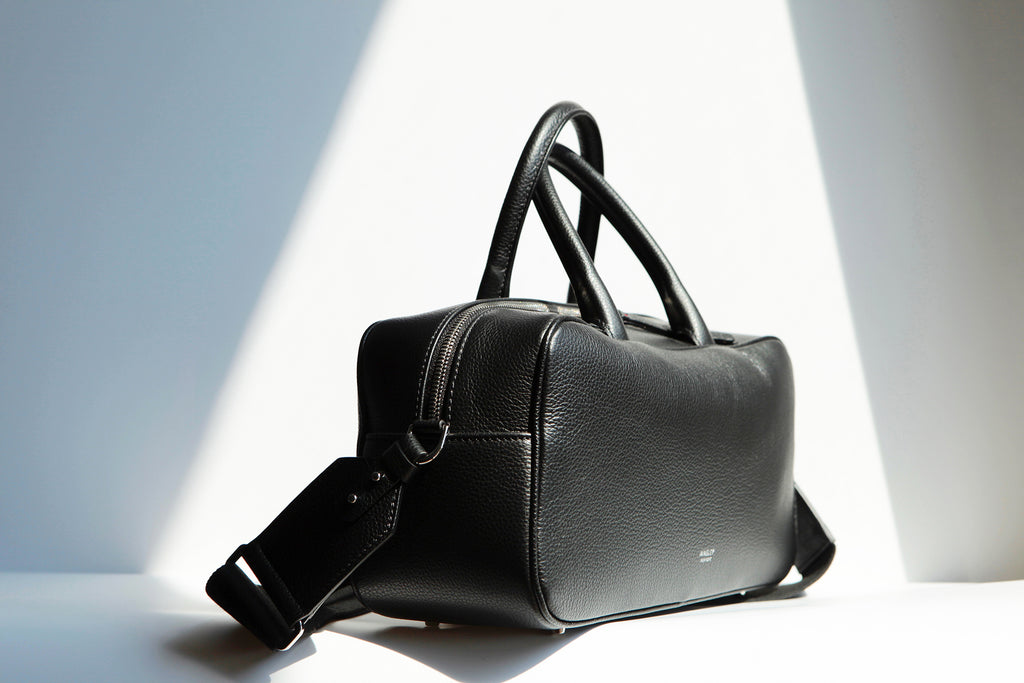 Side View of the Ines Mini Carryall Satchel. Shows the width and top zipper that extends on third way down the sides of the bag to allow for the top to open wide.  Bottom studs to protect the bottom leather corners.